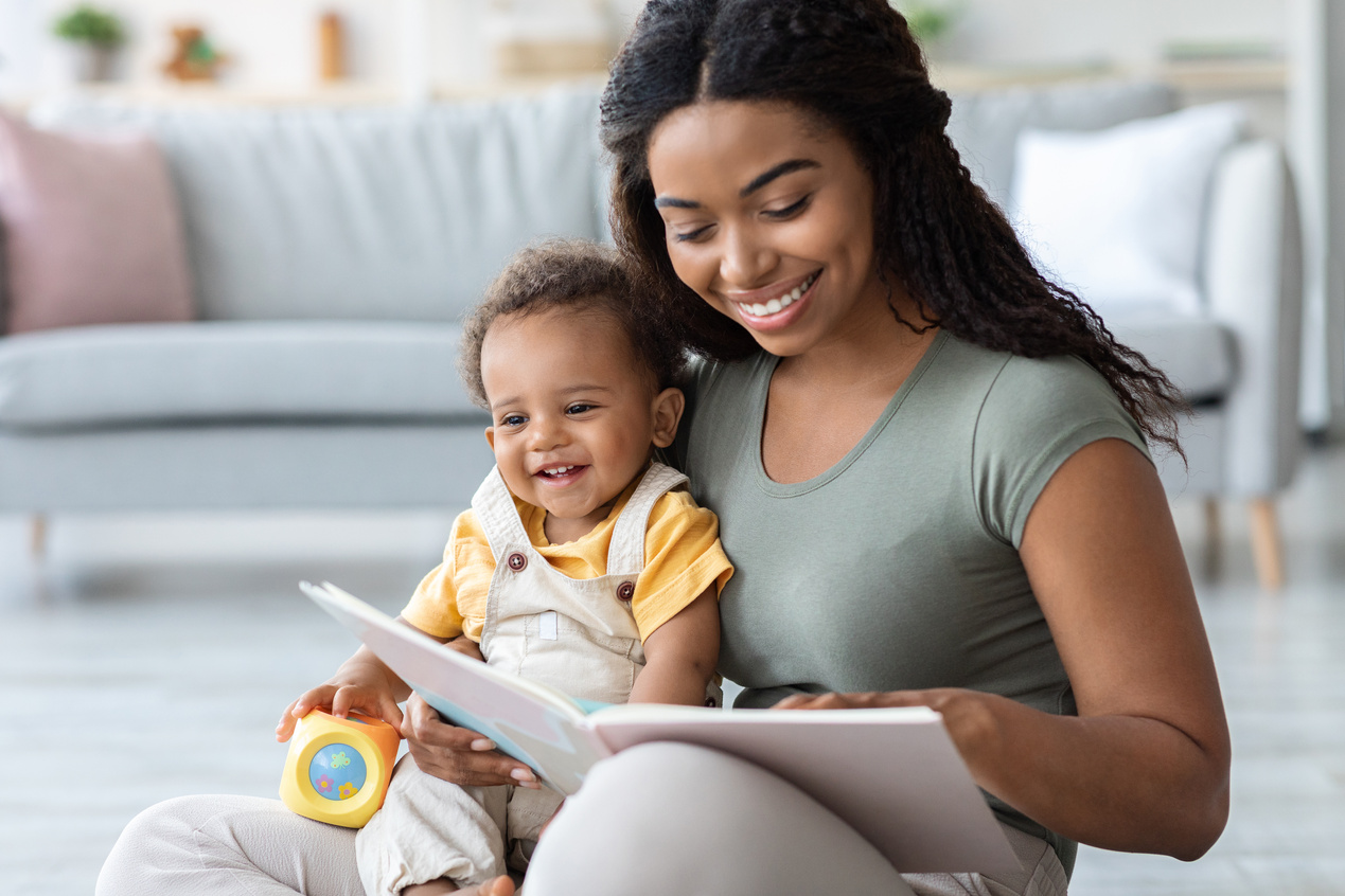 Baby Development. Young Black Mommy Reading Book with Infant Son at Home